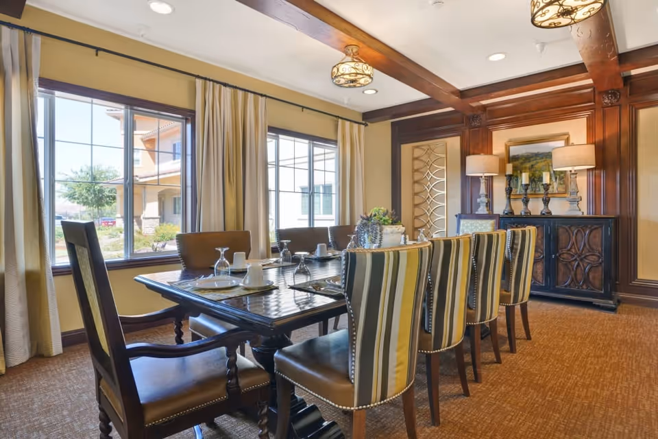 A well-lit dining room with a long rectangular table set for six people. The chairs have striped upholstery and nailhead trim. Large windows with beige curtains allow natural light to fill the room. The walls feature wood paneling and decorative elements, including a sideboard with two lamps and a framed painting above it.