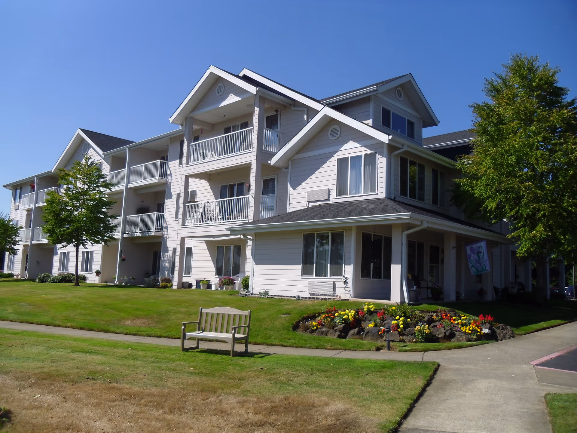 Exterior view of a three-story residential building with balconies, surrounded by a well-maintained lawn, trees, and a flower bed. A wooden bench is placed on the grass near a sidewalk under a clear blue sky.
