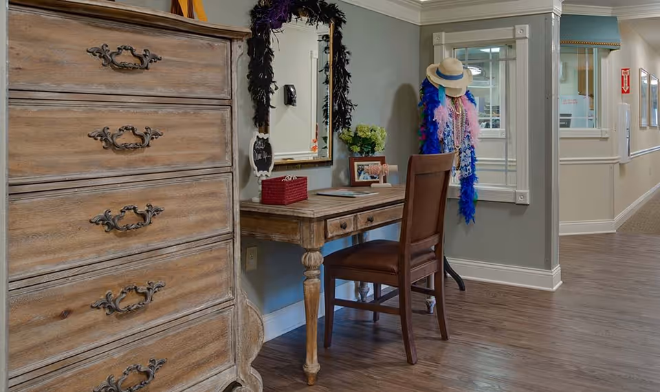 A cozy interior hallway area in a senior living facility featuring a wooden chest of drawers, a wooden desk with a chair, a mirror decorated with a black feather boa, and a coat rack holding hats and colorful feather boas. The hallway has wood flooring and light-colored walls with white trim.