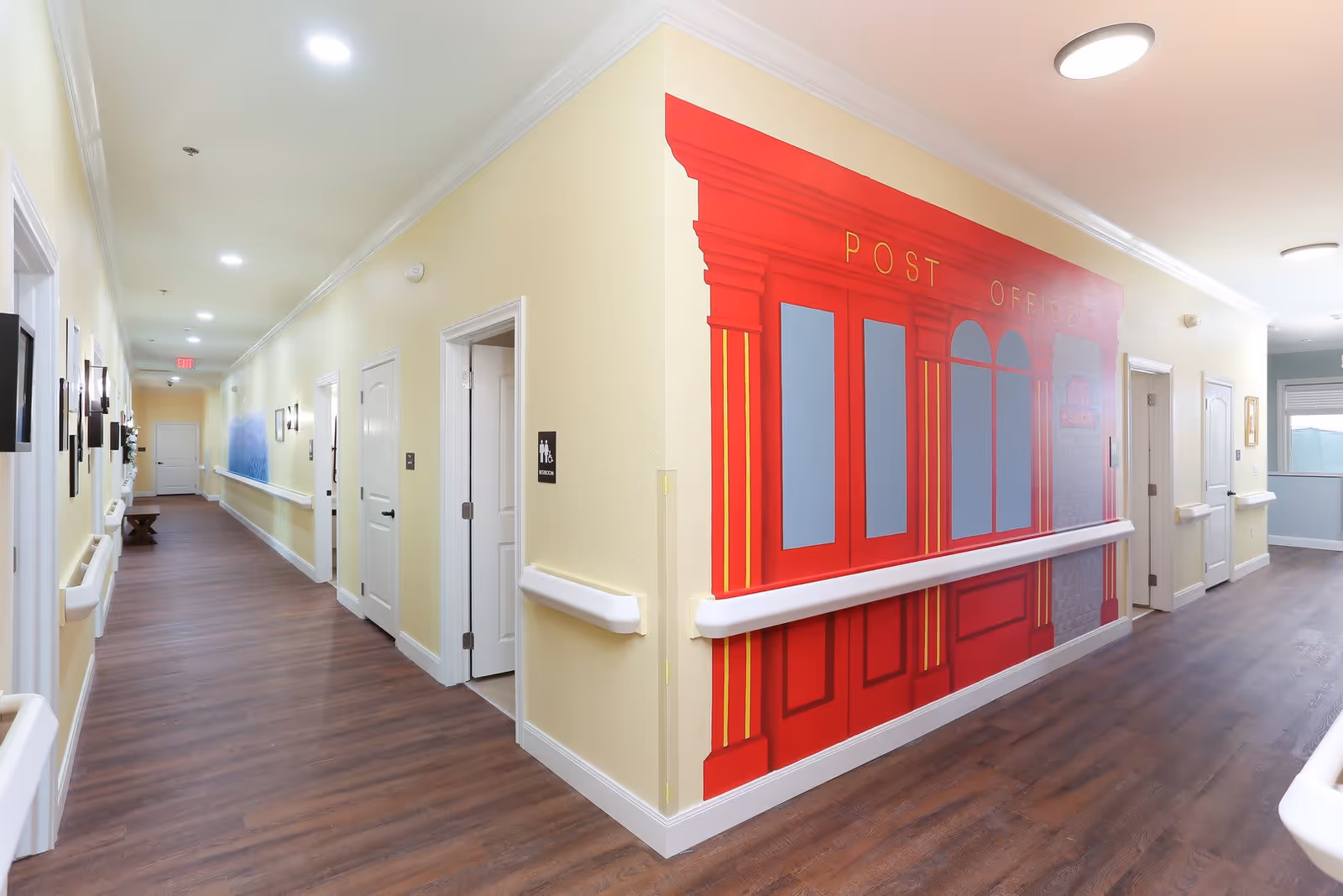 A brightly lit hallway in an assisted living facility with light yellow walls and wood flooring. The hallway features white doors on both sides, handrails along the walls, and a large red mural resembling a post office facade on one corner wall. There are ceiling lights and wall sconces providing illumination.