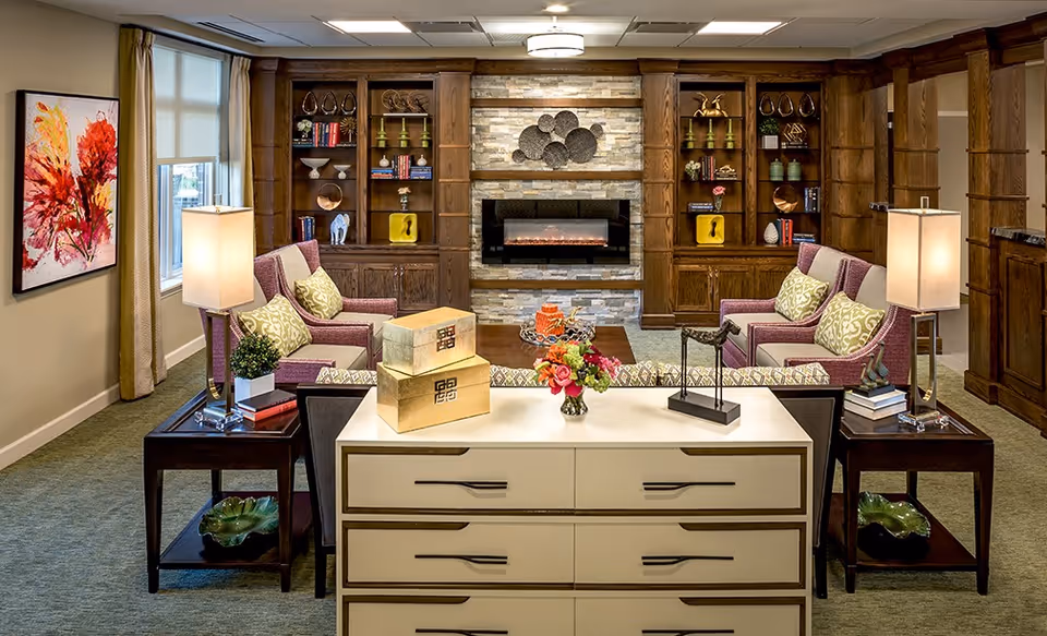 A cozy senior living room with two pairs of purple armchairs facing each other around a wooden coffee table. The room features built-in wooden bookshelves filled with decorative items and books, a modern electric fireplace with a stone surround, and two table lamps on dark wooden side tables. A colorful abstract painting hangs on the wall next to a window with beige curtains. A white dresser with decorative boxes and a flower vase is in the foreground.
