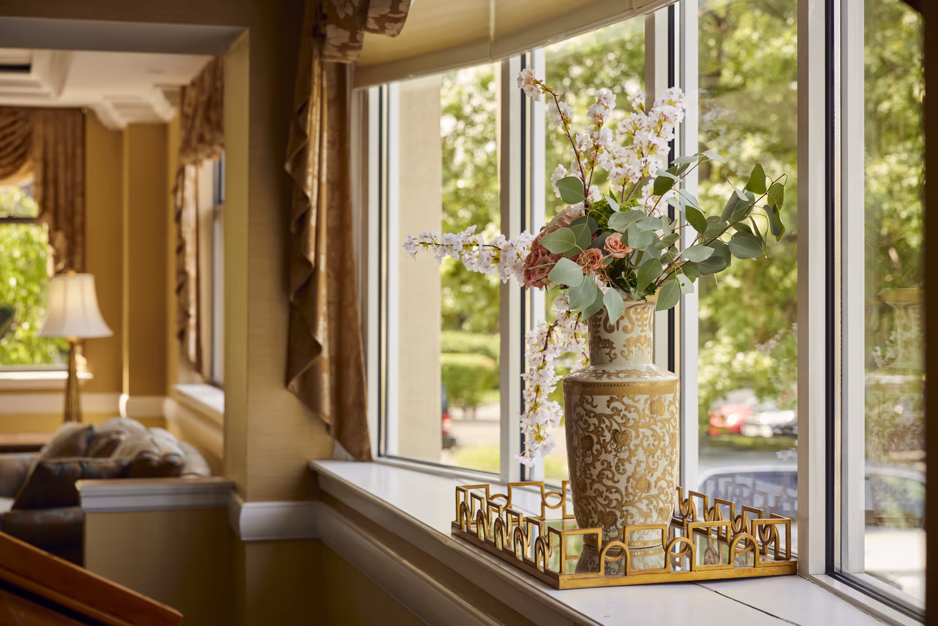 A decorative vase with intricate gold patterns holding a floral arrangement of white and pink flowers and green leaves sits on a gold-trimmed mirrored tray on a windowsill. The window overlooks a green outdoor area with trees and parked cars. Inside, the room features beige walls, gold curtains, and a lamp with a white shade near a cushioned seating area.