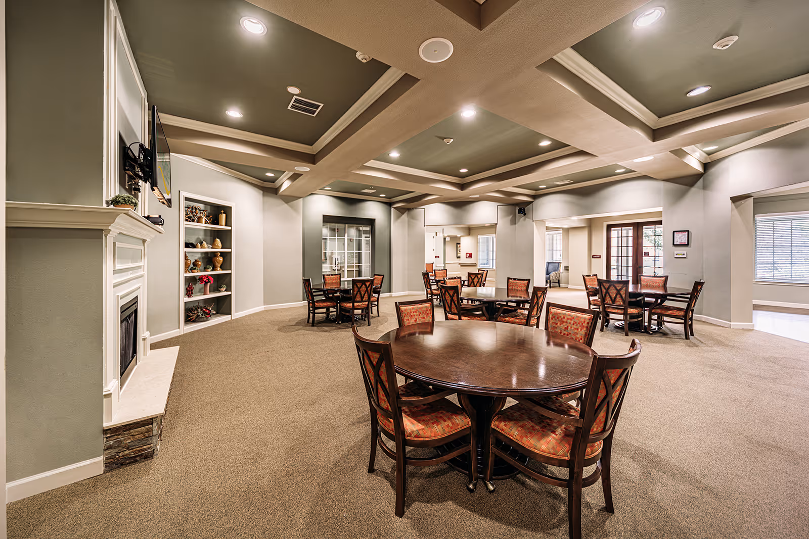 Spacious common area with multiple round wooden tables and chairs with patterned cushions, beige carpet, a white fireplace with a mounted TV above it, built-in shelves with decorative items, and a coffered ceiling with recessed lighting.