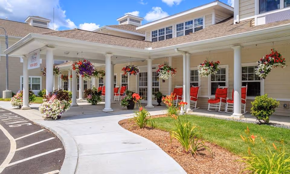 Front exterior view of All American Assisted Living at Hillsborough showing a covered entrance with white columns, hanging flower baskets, red cushioned rocking chairs, and landscaped garden beds with flowers under a partly cloudy blue sky.