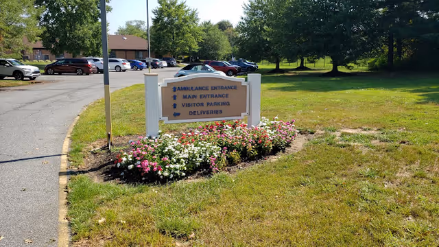 A landscaped entrance sign with directional text stands among flowers by a parking lot and grassy lawn.