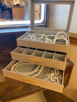 A beige jewelry box with two open drawers and a top compartment with a mirror, containing multiple strands of white pearls and pearl bracelets, placed on a wooden surface.