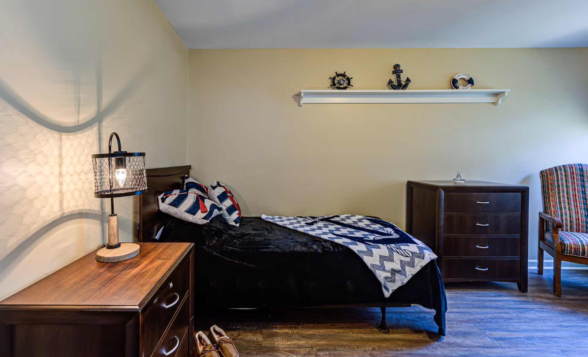 A cozy bedroom with a single bed covered in a black blanket and a chevron-patterned throw. Two pillows with nautical designs rest against the headboard. A wooden nightstand with a decorative lamp is beside the bed. On the wall above the bed, a white shelf holds nautical-themed decorations including a ship's wheel, an anchor, and a life preserver. To the right, there is a dark wooden dresser and a striped upholstered chair. The room has wooden flooring and light-colored walls.