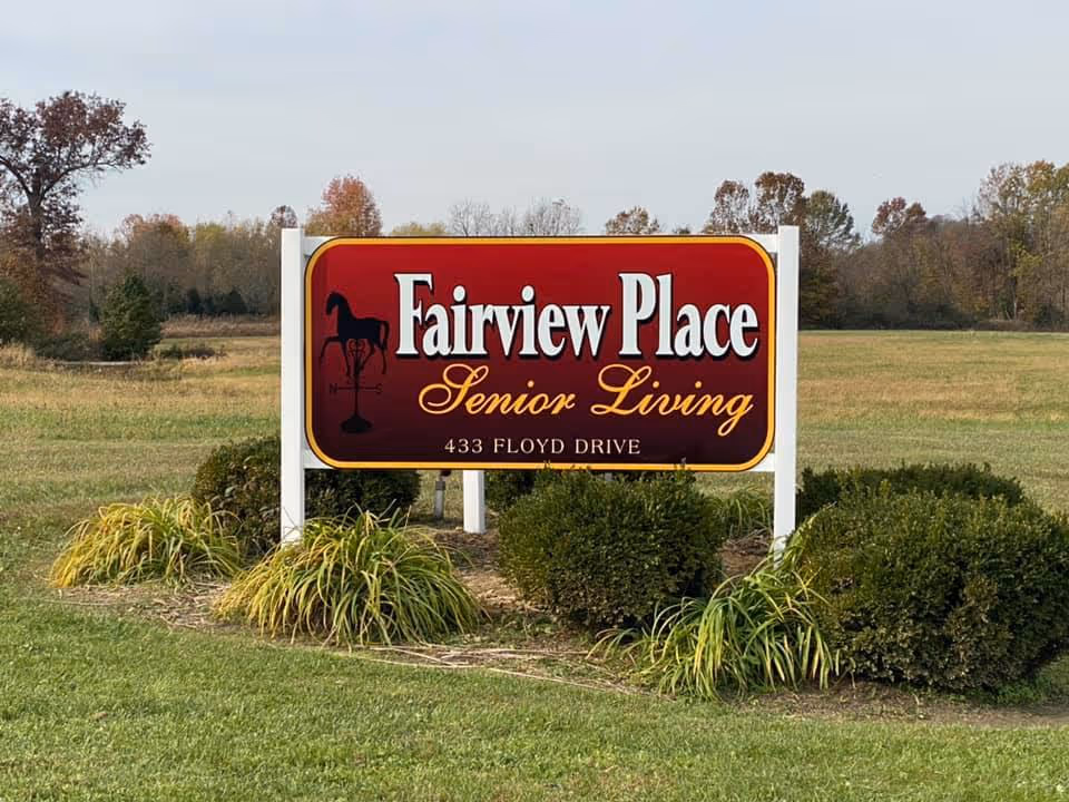 A large red and yellow sign for Fairview Place Senior Living at 433 Floyd Drive, set in a grassy area with bushes and trees in the background under a cloudy sky.