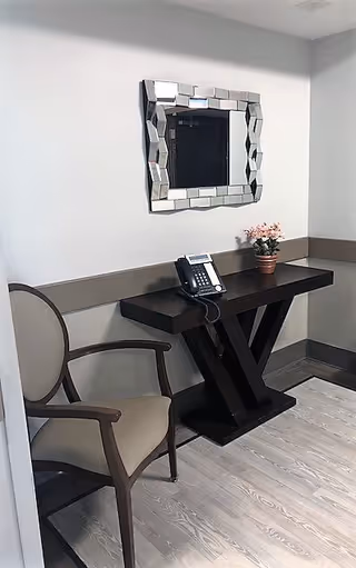 A small corner area with a wooden chair featuring beige upholstery, a dark wooden table with a unique X-shaped base, a telephone on the table, a small potted plant with pink flowers, and a decorative mirror with a geometric frame hanging on a light gray wall.