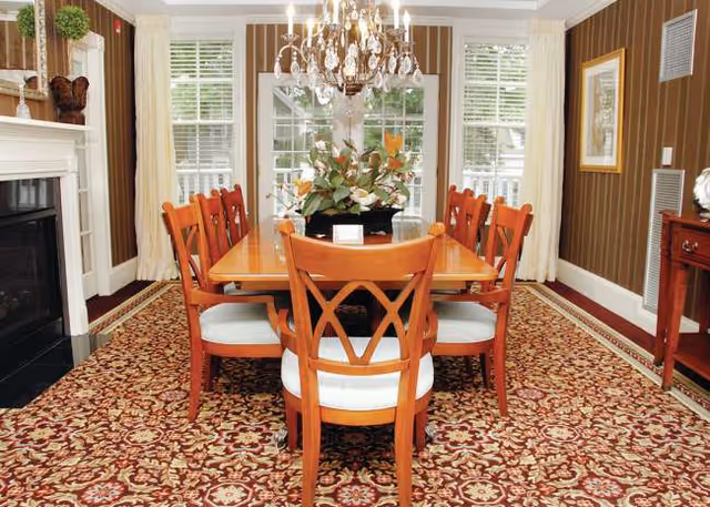 A formal dining room with a wooden table, red chairs, chandelier, and patterned carpet.