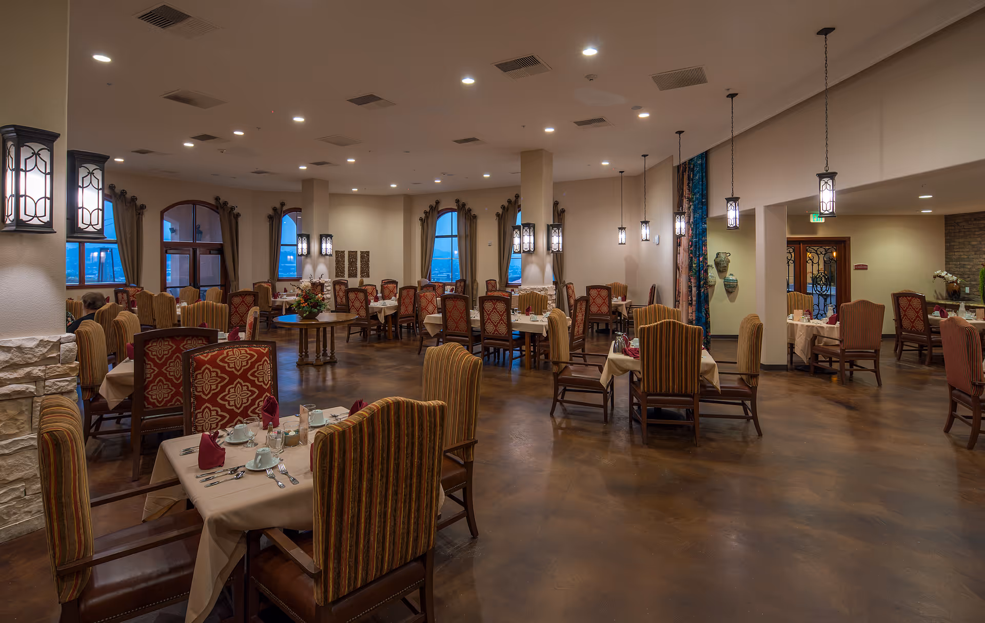 Spacious dining room in Lantern Crest Senior Living with multiple tables set with beige tablecloths, red napkins, and white dishware. The room features high ceilings with hanging lantern-style lights, large windows with curtains, and a polished brown floor. Chairs have patterned upholstery in red and gold tones.