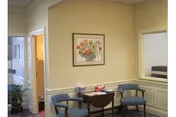 A small waiting area with two blue upholstered chairs and a wooden table between them. On the table are some tissues and a few small items. The walls are painted beige with white wainscoting. A framed floral painting hangs on the wall above the table. There is a doorway leading to another room on the left and a window opening on the right.