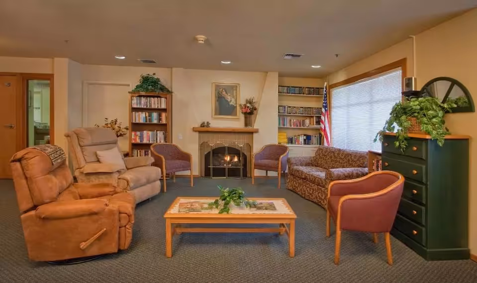 A cozy senior living common area with a fireplace in the center, flanked by bookshelves filled with books. The room has various seating options including two recliners, two purple armchairs, and a patterned sofa. A wooden coffee table with a plant on it is in the middle of the seating arrangement. There is a green chest of drawers with a plant on top near a window with blinds, and an American flag stands in the corner.