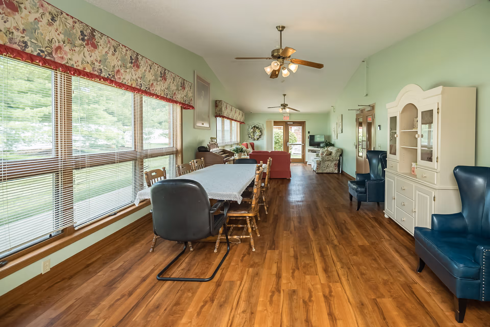 Sunlit common room with a long dining table and chairs, sofas, armchairs, and large windows in a senior living facility.