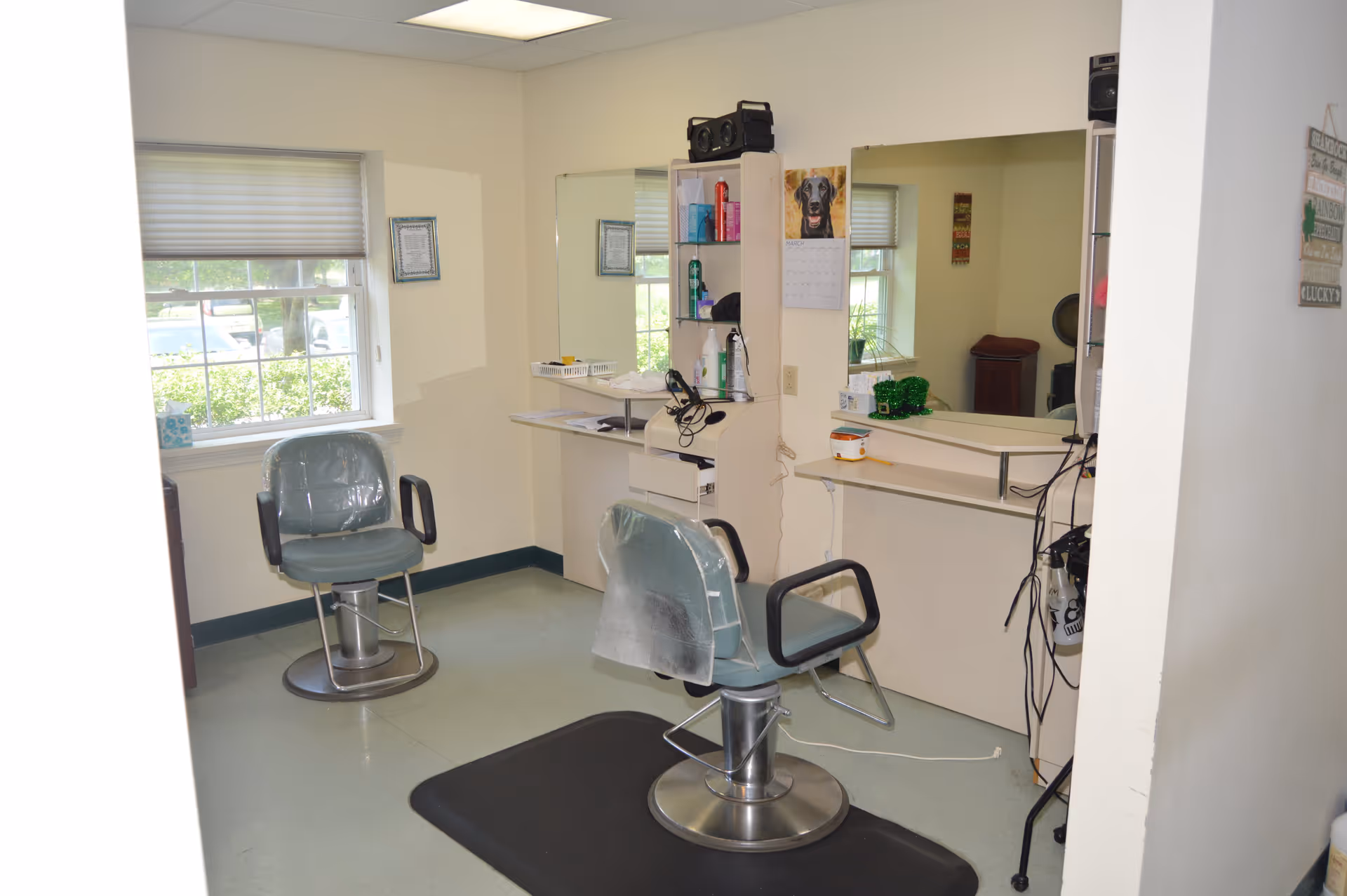 Interior view of a small salon or barber area with two green salon chairs, a large mirror on the wall, shelves with hair care products, and a window with blinds letting in natural light.