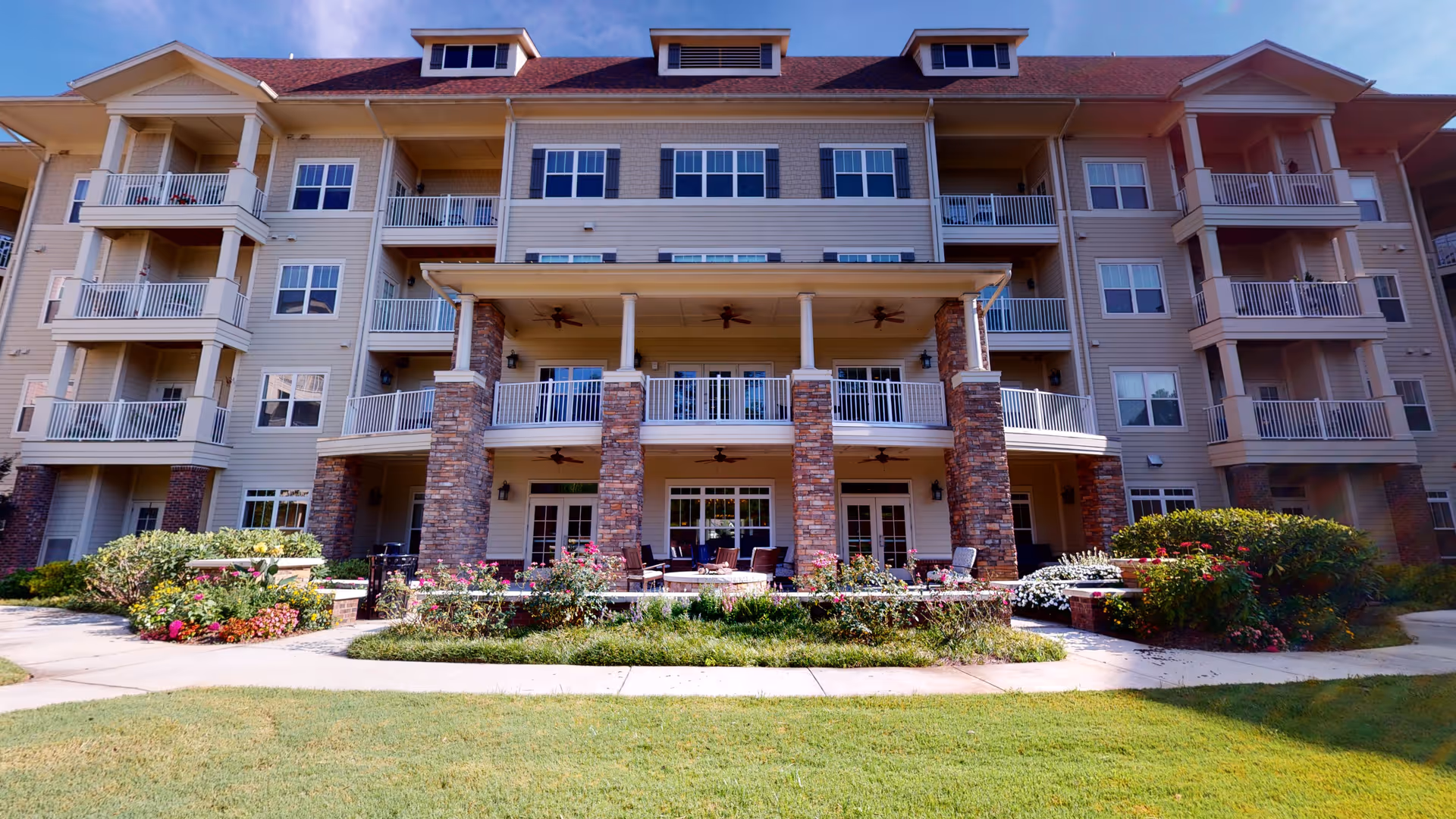 Front exterior of a multi-story senior living building with balconies, brick columns, patio seating, and landscaped lawn.