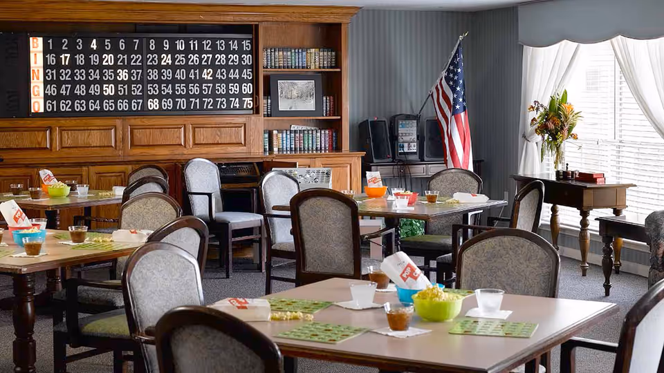 A senior living dining/activity room set up for bingo with tables and chairs, a large bingo board on a wood-paneled wall and an American flag by the window.