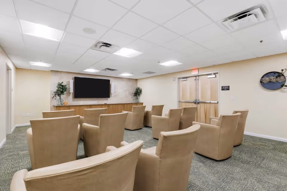 A small theater-style room with beige armchairs arranged in rows facing a large flat-screen TV mounted on a wall. The room has a drop ceiling with fluorescent lights, light-colored walls, green patterned carpet, and double doors at the back. There are decorative plants on either side of the TV and a wall decoration on the right side.