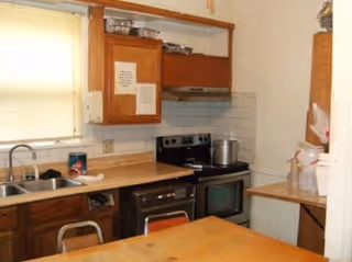Cozy facility kitchen with cabinets, stove, sink, and a wooden table.