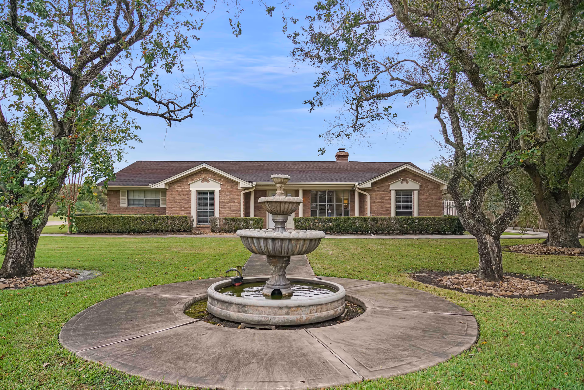 Front view of a brick memory care facility with a fountain, trees, and manicured lawn.