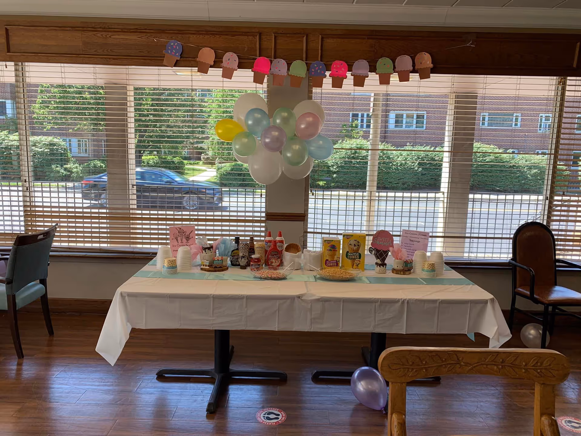 A decorated table set up for an ice cream social event inside a room with large windows and wooden blinds. The table is covered with a white tablecloth and has various ice cream toppings, cones, cups, and utensils. Above the table, there are pastel-colored balloons and a garland of paper ice cream cones. Chairs are placed around the room, and outside the window, a car and greenery are visible.