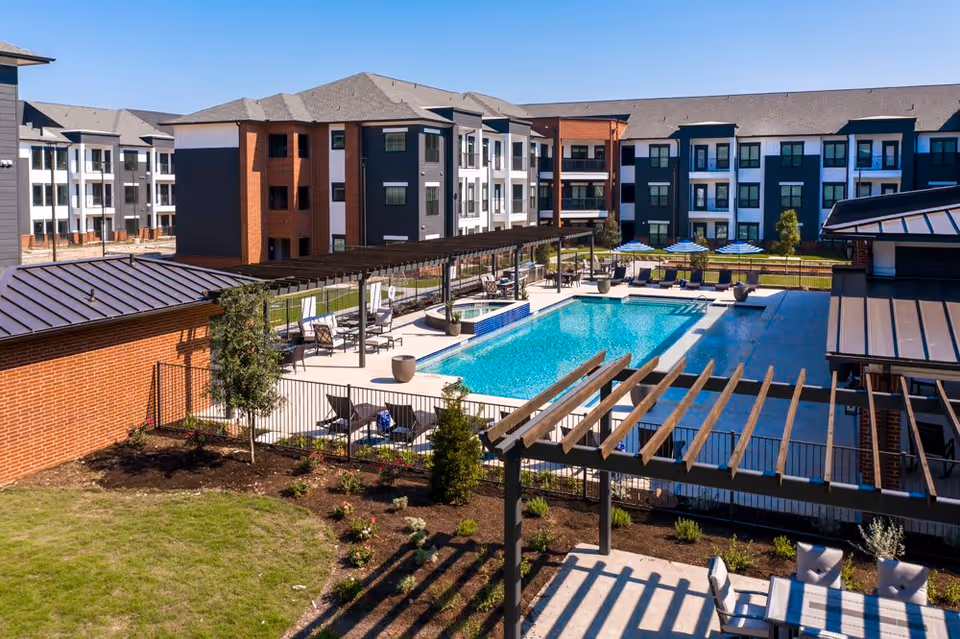 Outdoor view of NorthStar Georgetown senior living facility showing a large swimming pool surrounded by lounge chairs and umbrellas. The pool area is enclosed by a fence and features pergolas with seating areas. In the background, there are multi-story residential buildings with balconies and windows.