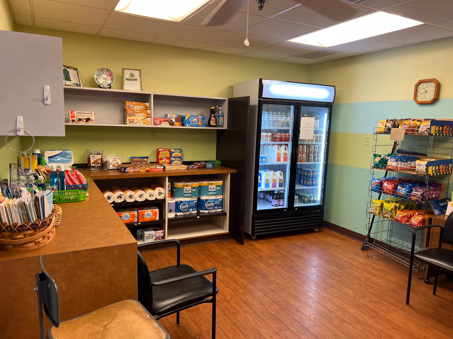 A small convenience store area inside a facility with shelves stocked with snacks, paper towels, and other supplies. There is a refrigerated display case filled with beverages including milk, soda, and water. A rack on the right holds various bags of chips. Two chairs are placed near the counter, and the walls are painted in light green and blue stripes with a clock hanging on the wall.