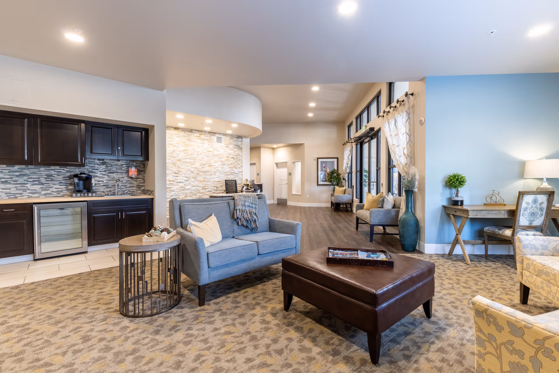 A bright and spacious living room area in Mukilteo Memory Care featuring a blue loveseat with pillows and a throw blanket, a round side table, a large brown leather ottoman with a tray on top, patterned carpet, and a kitchenette with dark cabinets and a wine cooler. The room has large windows with sheer curtains, a blue accent wall with a desk and chair, and additional seating along the hallway.