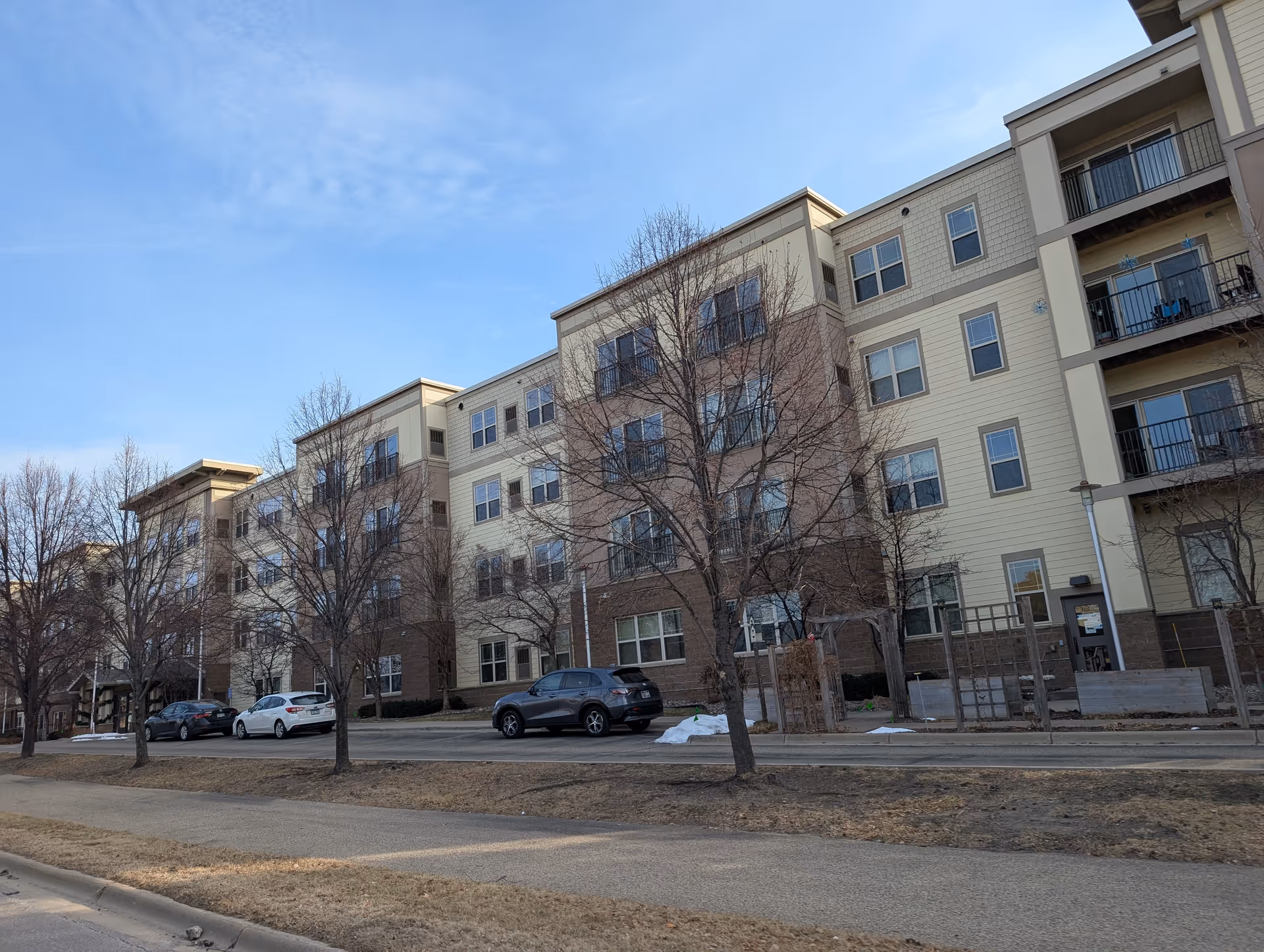 Front exterior of a four-story beige and brick senior living building with balconies, parked cars, and leafless trees along the street.