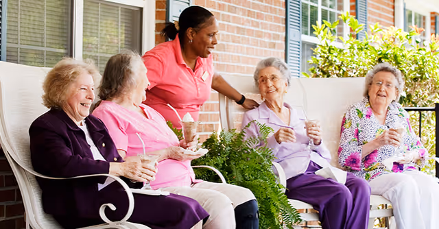 Four elderly women sitting on a porch swing and chairs, enjoying drinks and smiling while a caregiver in a pink uniform stands behind them, engaging in conversation. The setting is outdoors with a brick wall and greenery in the background.