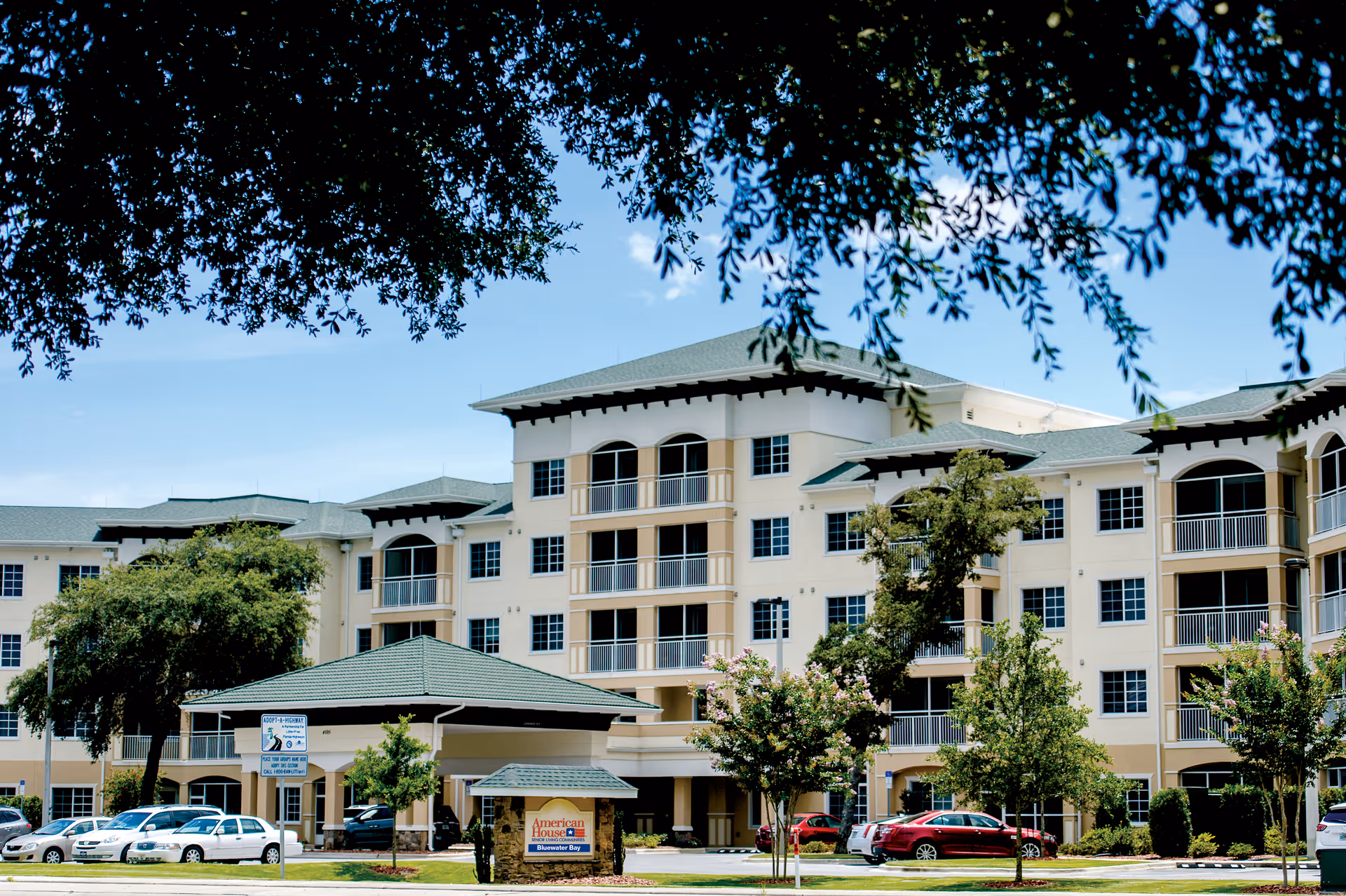 Exterior view of a multi-story senior living facility named American House Bluewater Bay, with a covered entrance, several parked cars, and trees framing the scene under a clear blue sky.