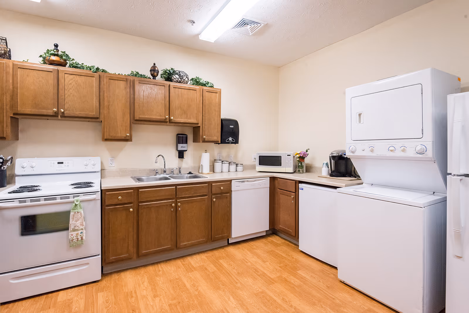 A clean communal kitchen with wooden cabinets, a stove, sink, microwave, dishwasher, and a stacked washer-dryer.