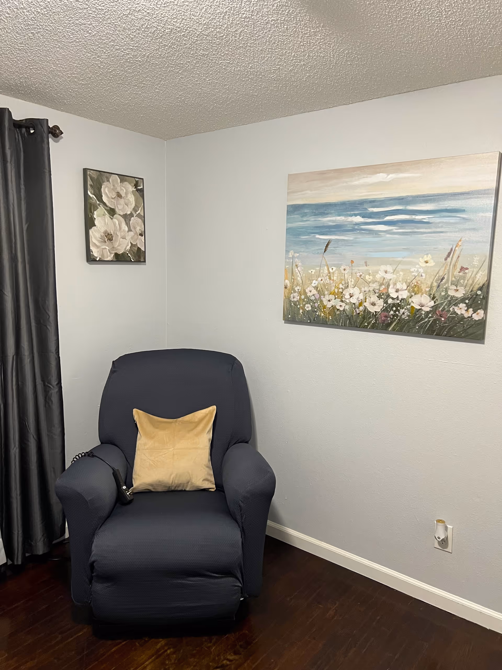 A cozy corner of a room with a dark blue recliner chair covered with a beige pillow. The walls are light gray with two floral paintings hanging on them. One painting depicts white flowers on a green background, and the other shows a field of flowers with a blue sky and ocean in the background. There is a dark curtain on the left side and a dark wooden floor.