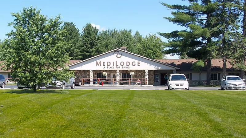 Front exterior of the Medilodge building with a wide green lawn, trees, parked cars, and an entrance canopy reading 'Medilodge A Place for Living'.