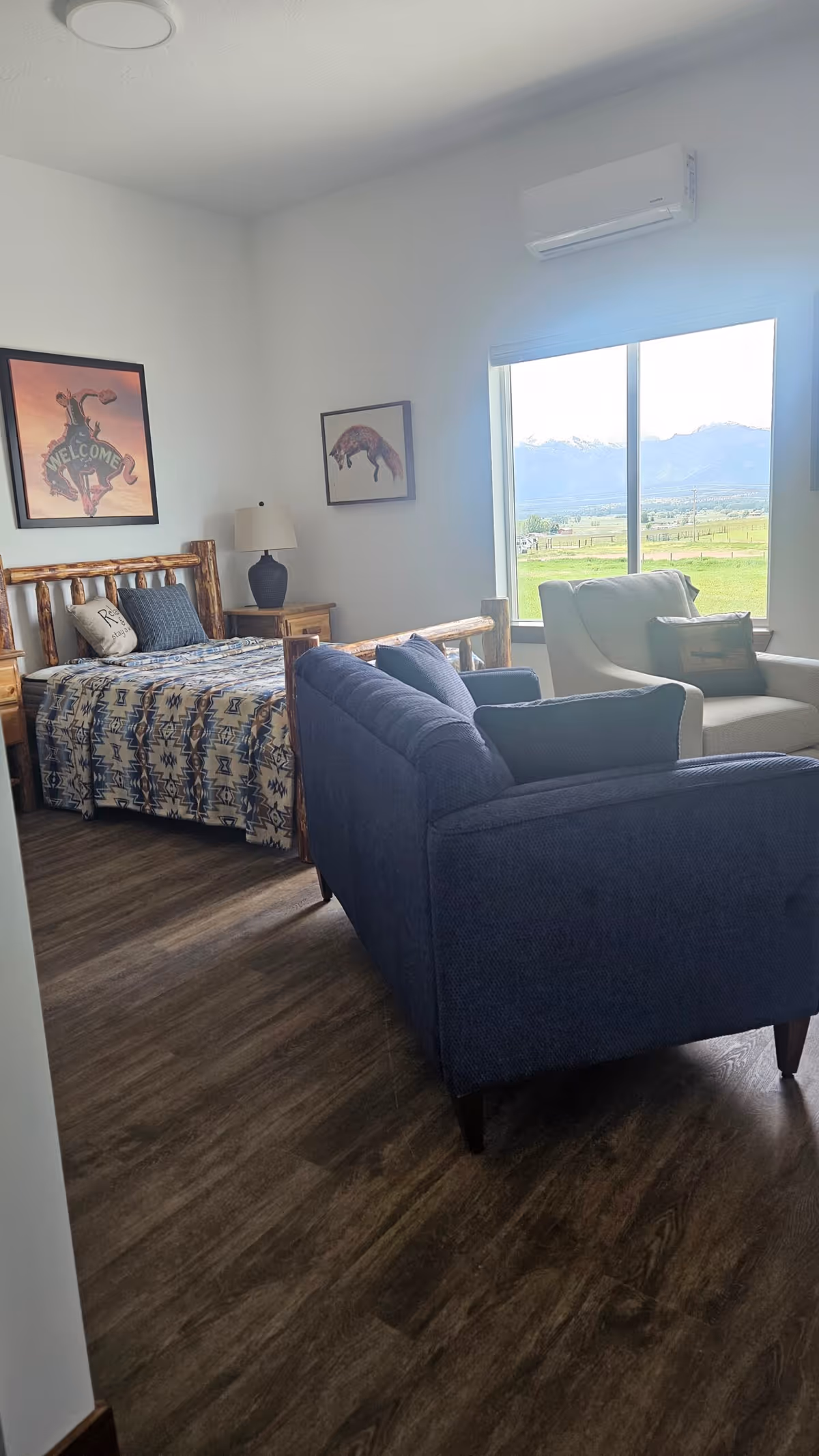 A cozy senior living bedroom featuring a wooden bed with patterned bedding, a nightstand with a lamp, a blue upholstered sofa, and a beige armchair near a large window with a scenic view of green fields and mountains. The walls have two framed artworks, and there is a wall-mounted air conditioning unit above the window.