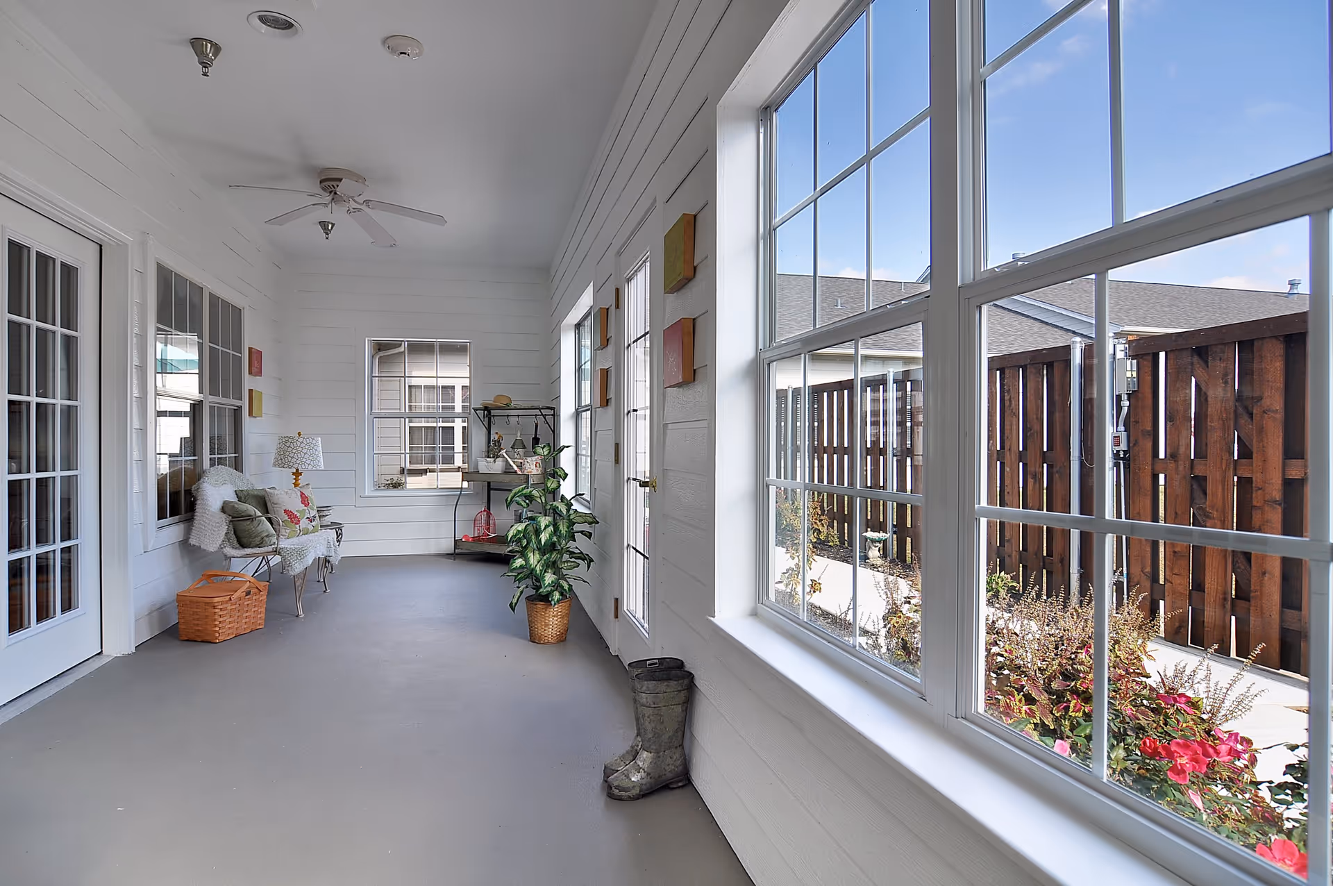 A bright enclosed porch area with large windows on the right side showing a view of a wooden fence and garden outside. Inside, there is a white bench with cushions and a throw blanket, a wicker basket, a potted plant, a small shelving unit with decorative items, and a ceiling fan overhead. The walls are white with horizontal paneling.