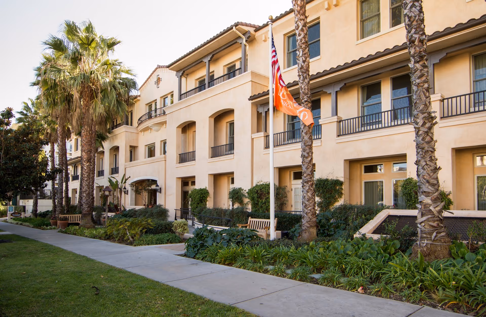 Exterior front of a multi-story beige senior living building with palm trees, landscaping, benches, and an American flag.