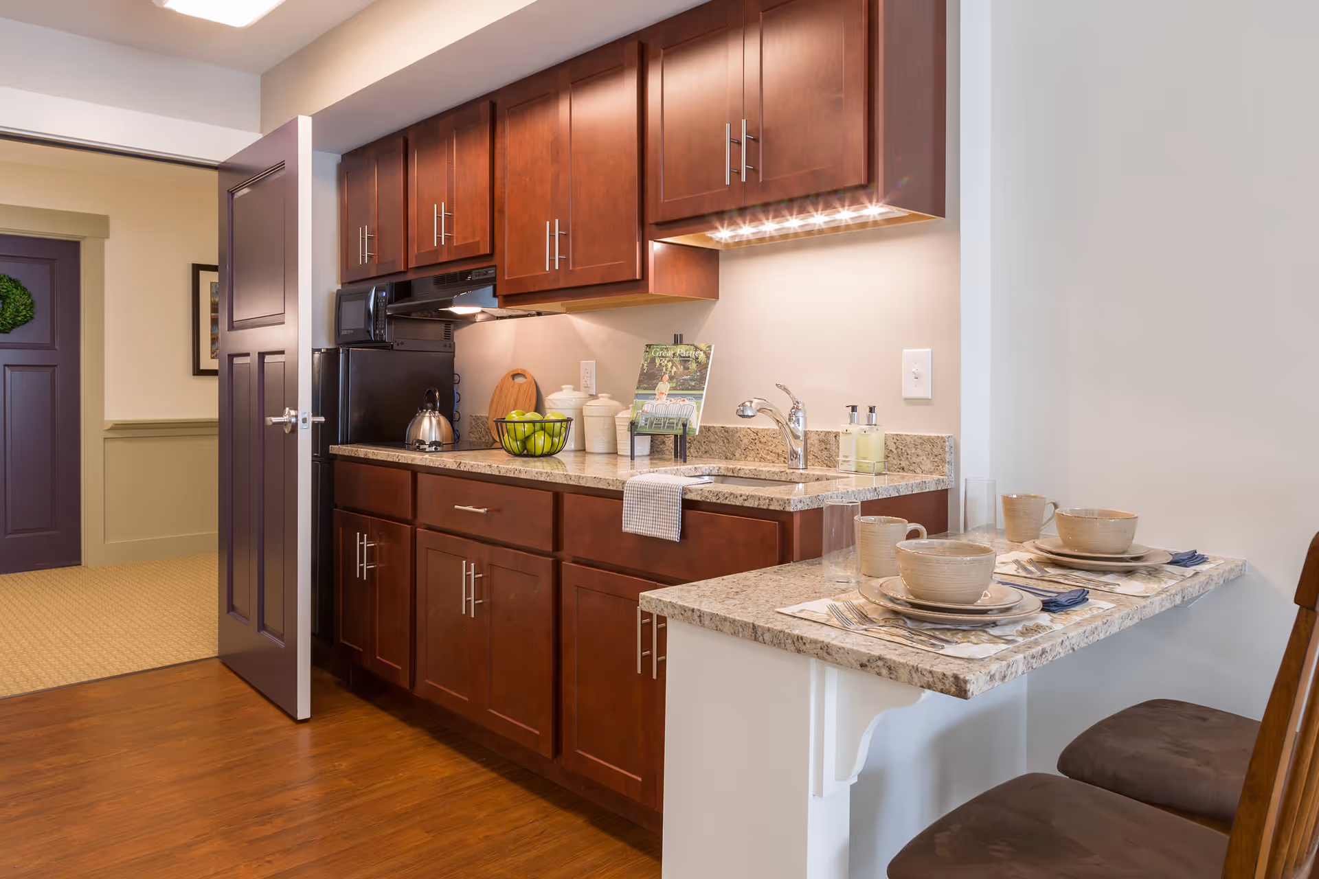 A modern kitchen area with dark wooden cabinets, granite countertops, a sink, and a small breakfast bar set with two place settings including bowls, plates, mugs, and utensils. The kitchen includes a microwave, a kettle, and decorative items such as a bowl of green apples and canisters. The floor is wooden, and there is an open door leading to a hallway with a purple door and green trim.