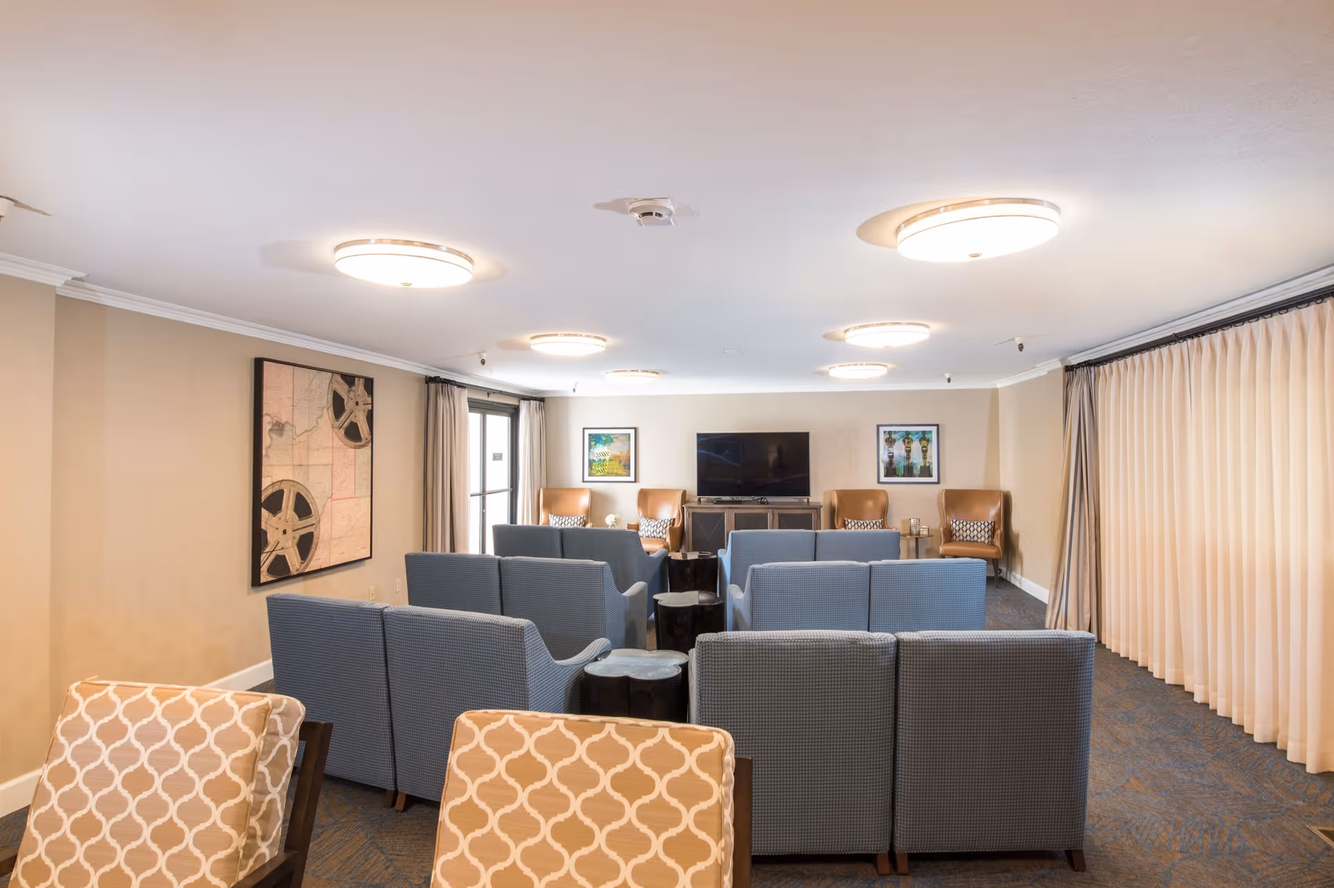A well-lit common living area with multiple rows of blue cushioned chairs facing a flat-screen TV mounted on a wooden cabinet. The room has beige walls, ceiling lights, framed artwork on the walls, and large curtains covering windows on the right side.