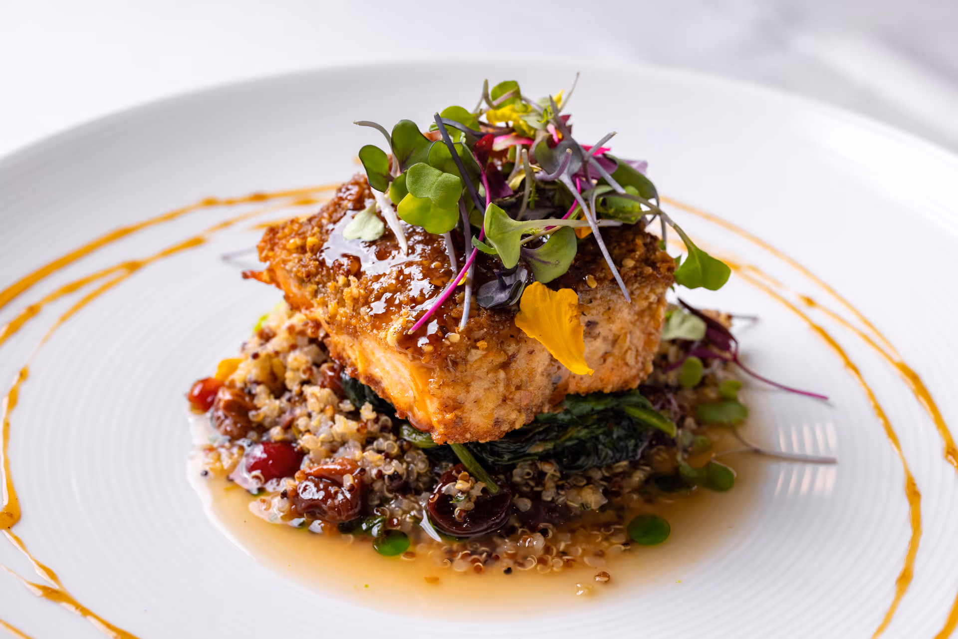 A gourmet dish featuring a piece of crusted salmon topped with microgreens and edible flowers, served on a bed of quinoa and sautéed greens, with a decorative sauce drizzle on a white plate.