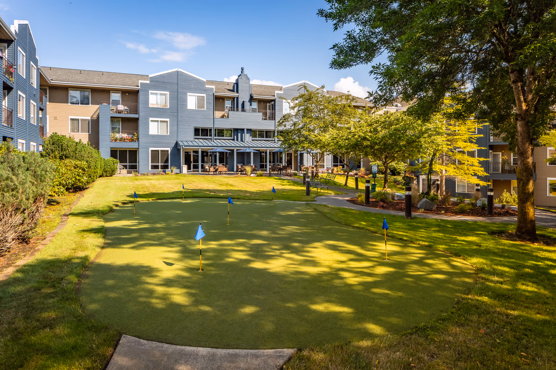 A courtyard putting green with flags in front of a three-story senior living building surrounded by trees.