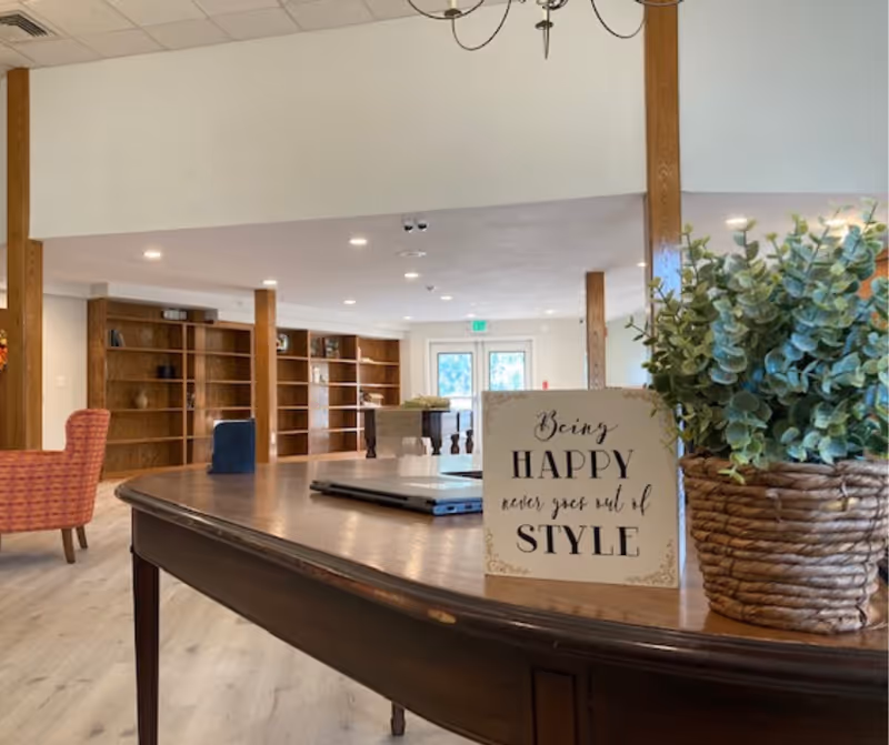 Bright communal lounge with a wooden table in the foreground holding a potted plant and decorative sign, with shelving and chairs in the background.