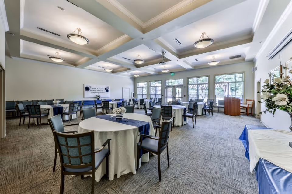 A spacious dining room with multiple round tables covered with white tablecloths and blue runners, surrounded by chairs. The room features large windows allowing natural light to fill the space, a coffered ceiling with modern light fixtures, and a carpeted floor. There are floral centerpieces on the tables and a podium near the windows.