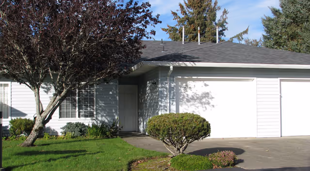 Exterior view of a single-story residential building with white siding, a gray roof, two white garage doors, a front door, and a small landscaped area with green grass, bushes, and a tree with dark red leaves.