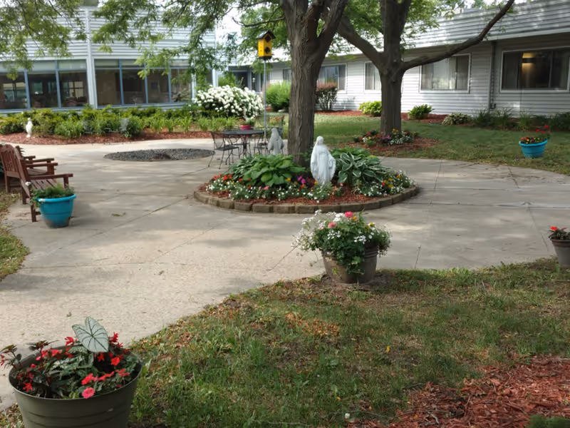 Outdoor courtyard area with a circular flower bed containing a statue and plants, surrounded by trees and paved walkways. There are several potted plants with flowers placed around the courtyard, wooden benches, and a metal table with chairs. The building with windows and white siding is visible in the background.