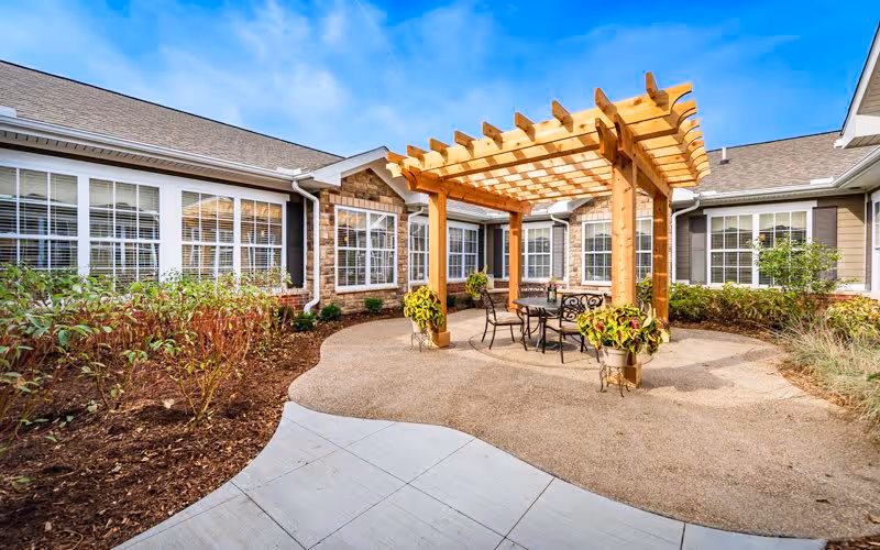 Outdoor courtyard area at Bickford of Shelby Township featuring a wooden pergola with a table and chairs underneath, surrounded by plants and shrubs, with building windows in the background under a blue sky.