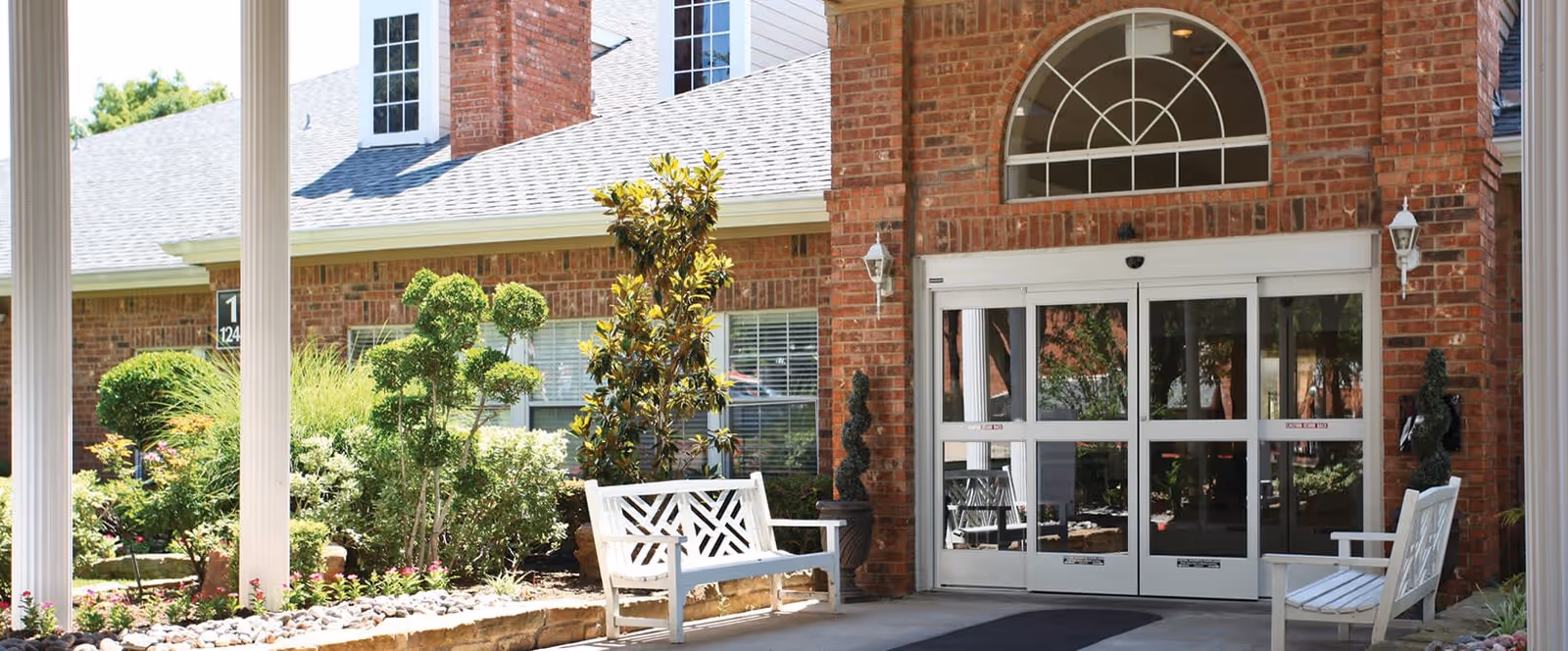 Entrance of a senior living facility with a brick exterior, white framed glass double doors, two white benches on either side of the entrance, and well-maintained landscaping including bushes and small trees.