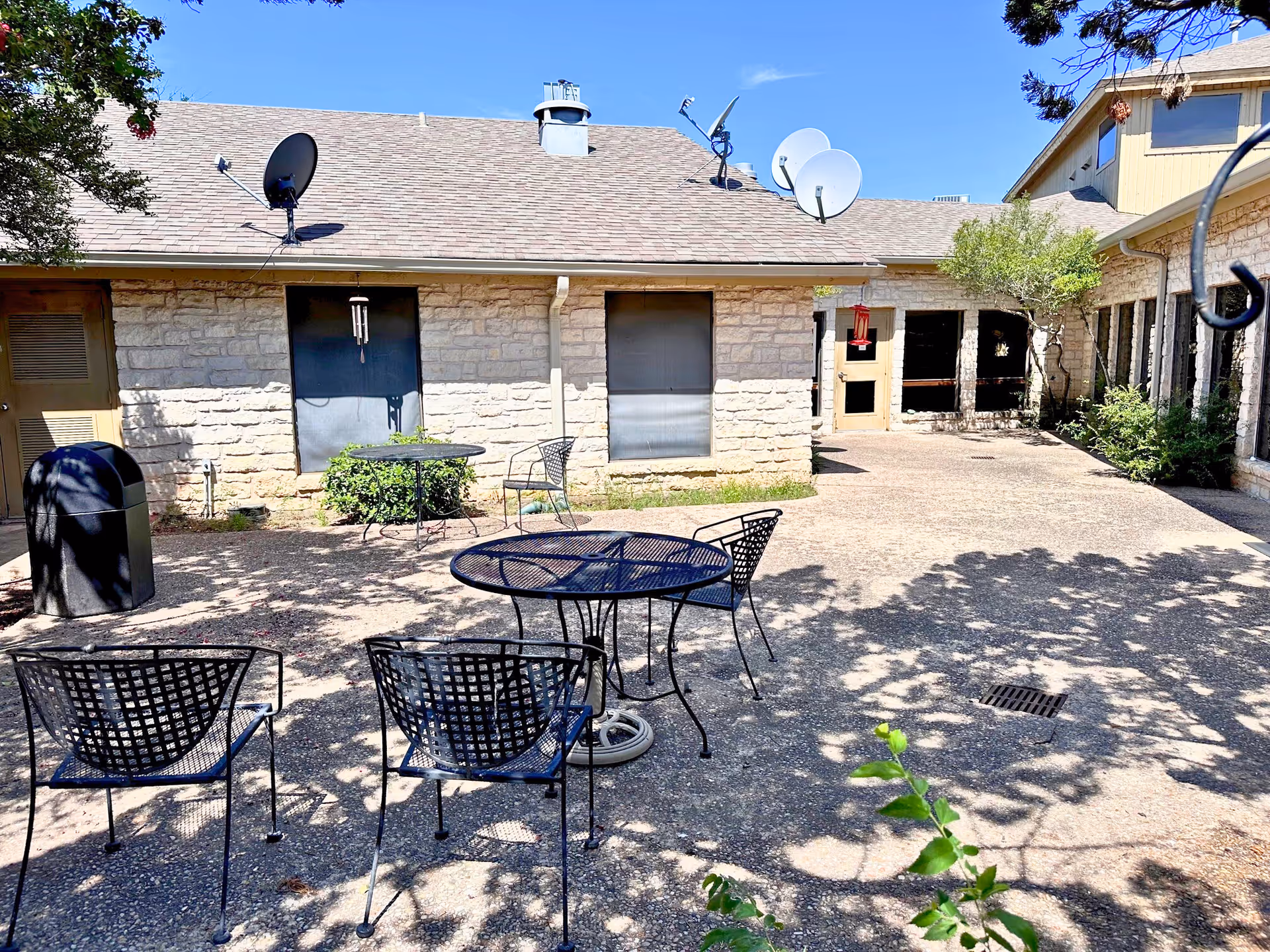 Courtyard patio with metal tables and chairs in front of a low stone building with satellite dishes on the roof.