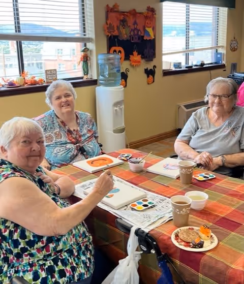 Three elderly women sitting around a table covered with a colorful checkered tablecloth, engaging in a painting activity with watercolor palettes and brushes. The table also has cups of tea and a plate with snacks. The room has large windows with blinds, a water cooler, and autumn-themed decorations on the wall.