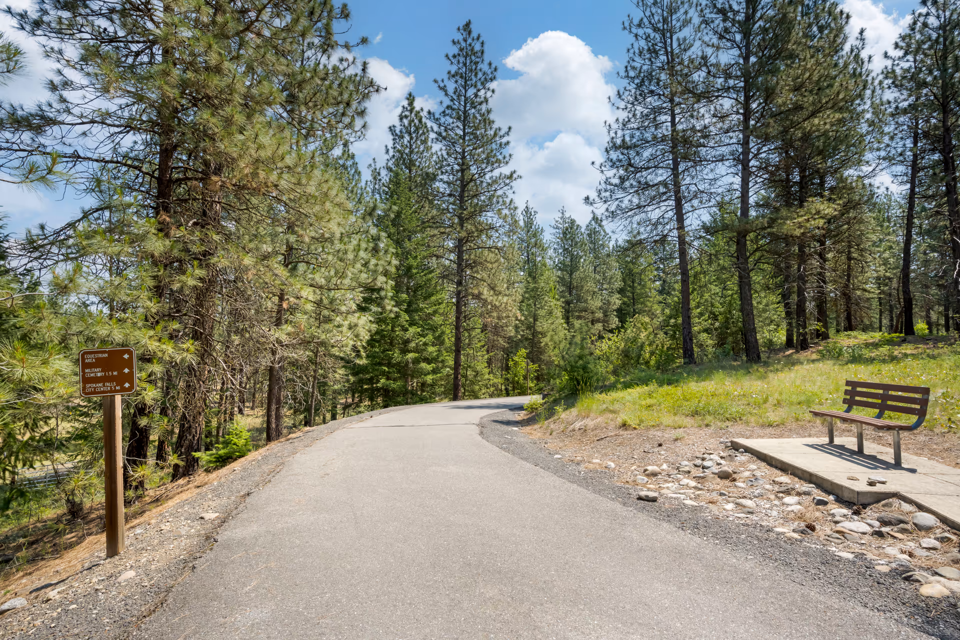A paved walking path winding through a forested area with tall pine trees under a partly cloudy sky. There is a wooden bench on a concrete slab to the right of the path and a brown signpost on the left side indicating directions to an equestrian area, military cemetery, and Spokane Falls city center.